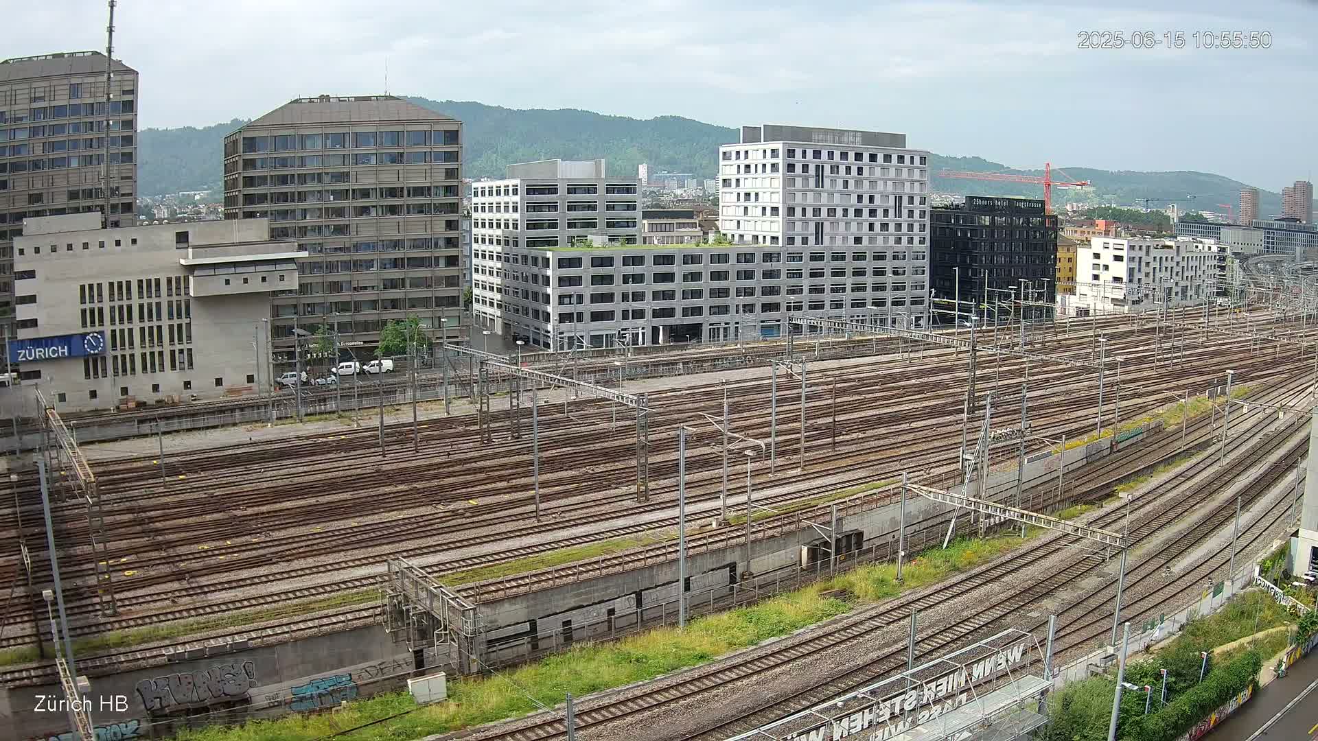 An expansive railway yard with numerous tracks stretches out before a backdrop of modern buildings under a partly cloudy sky.