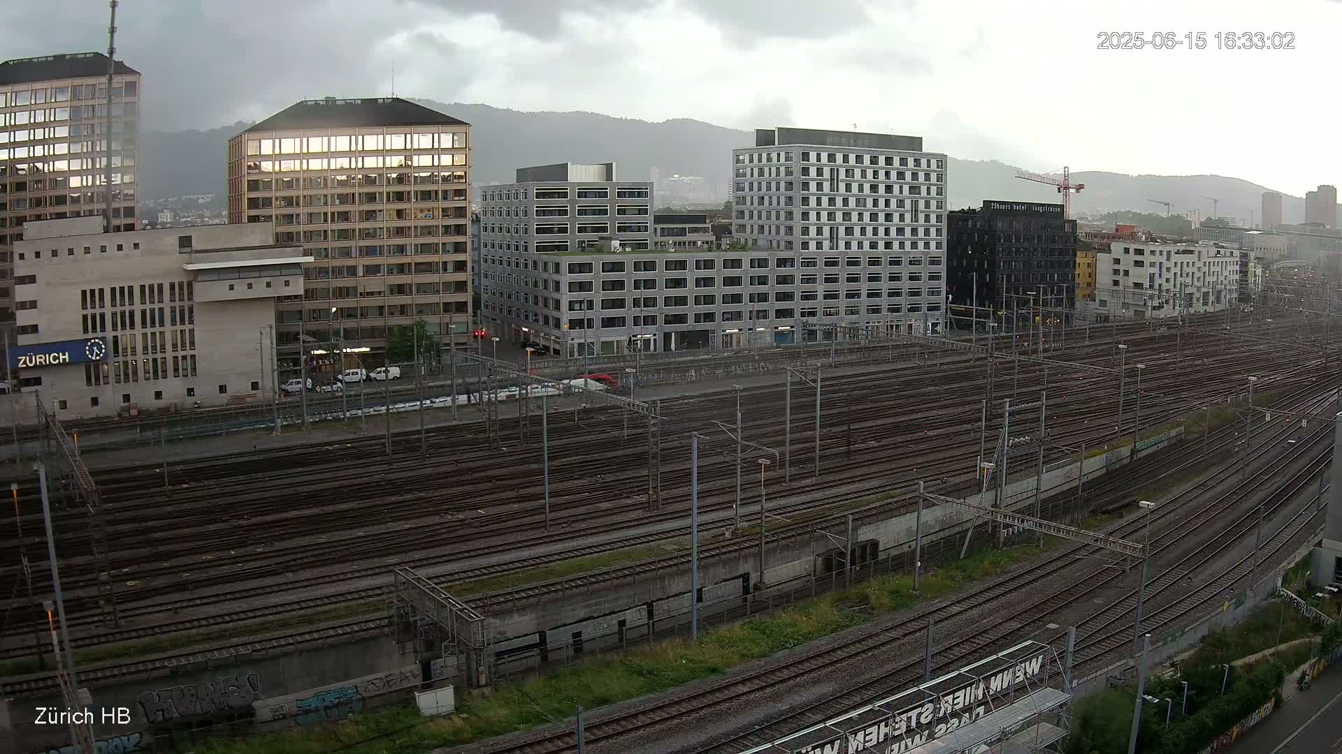 A sprawling railway yard with numerous train tracks stretches before a cityscape of modern buildings under a cloudy sky.