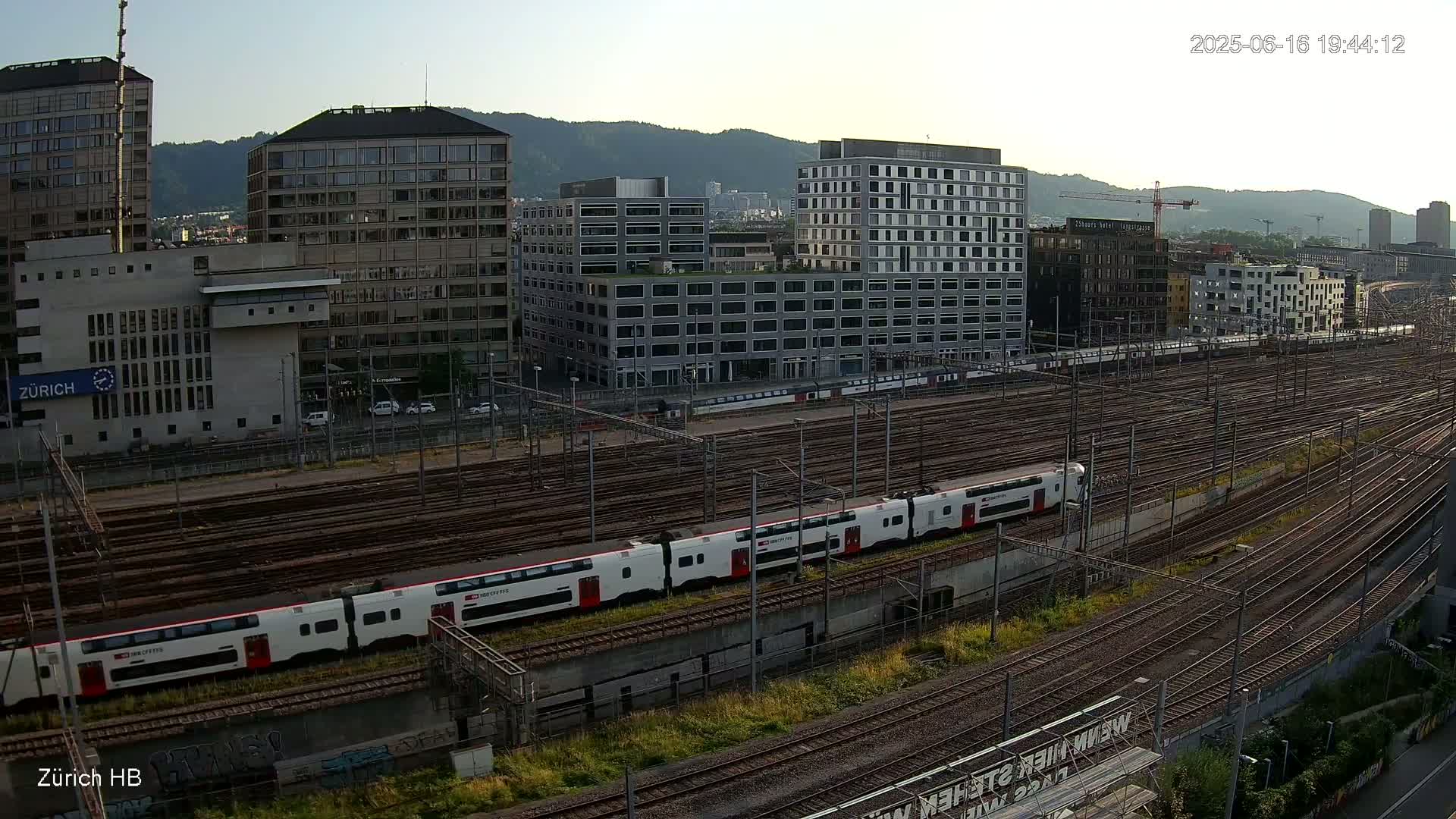 A long white and red train travels across a vast network of train tracks surrounded by modern buildings under a clear sky.