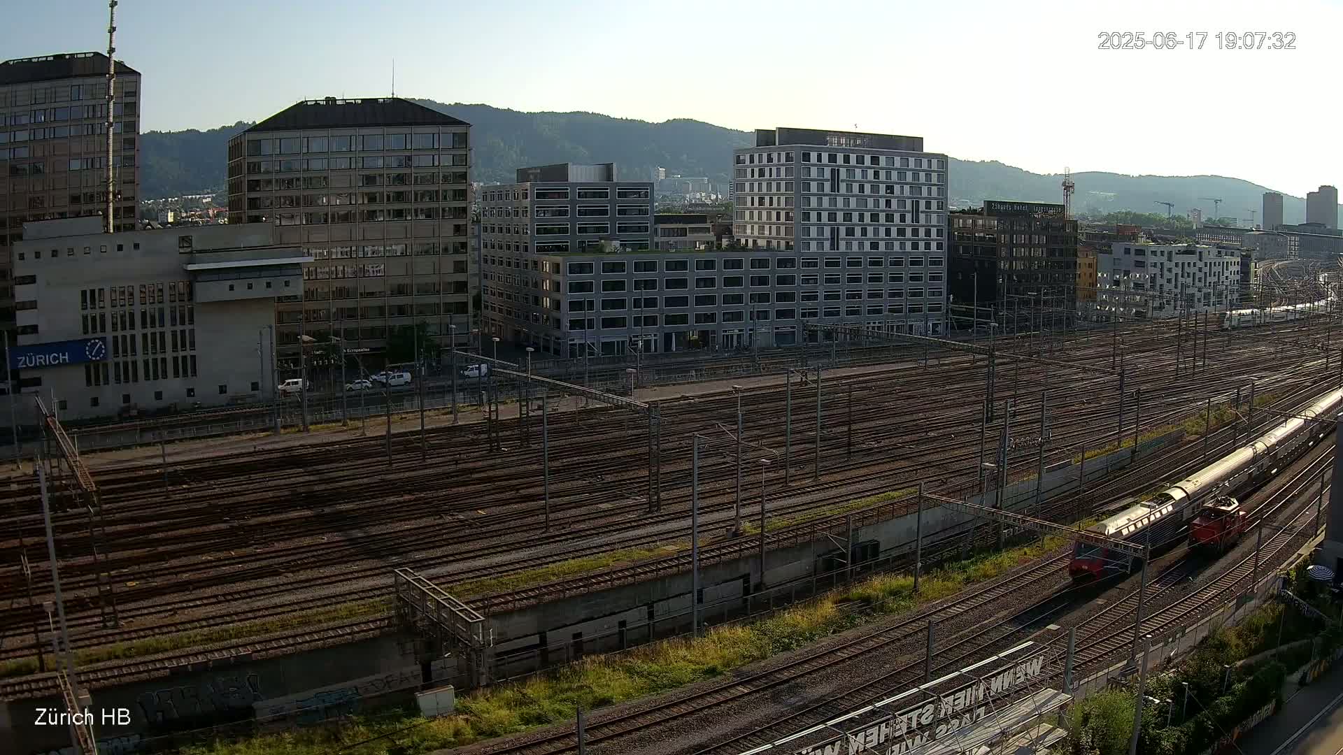 A long train passes through a large railway yard next to several modern and older buildings under a clear sky.