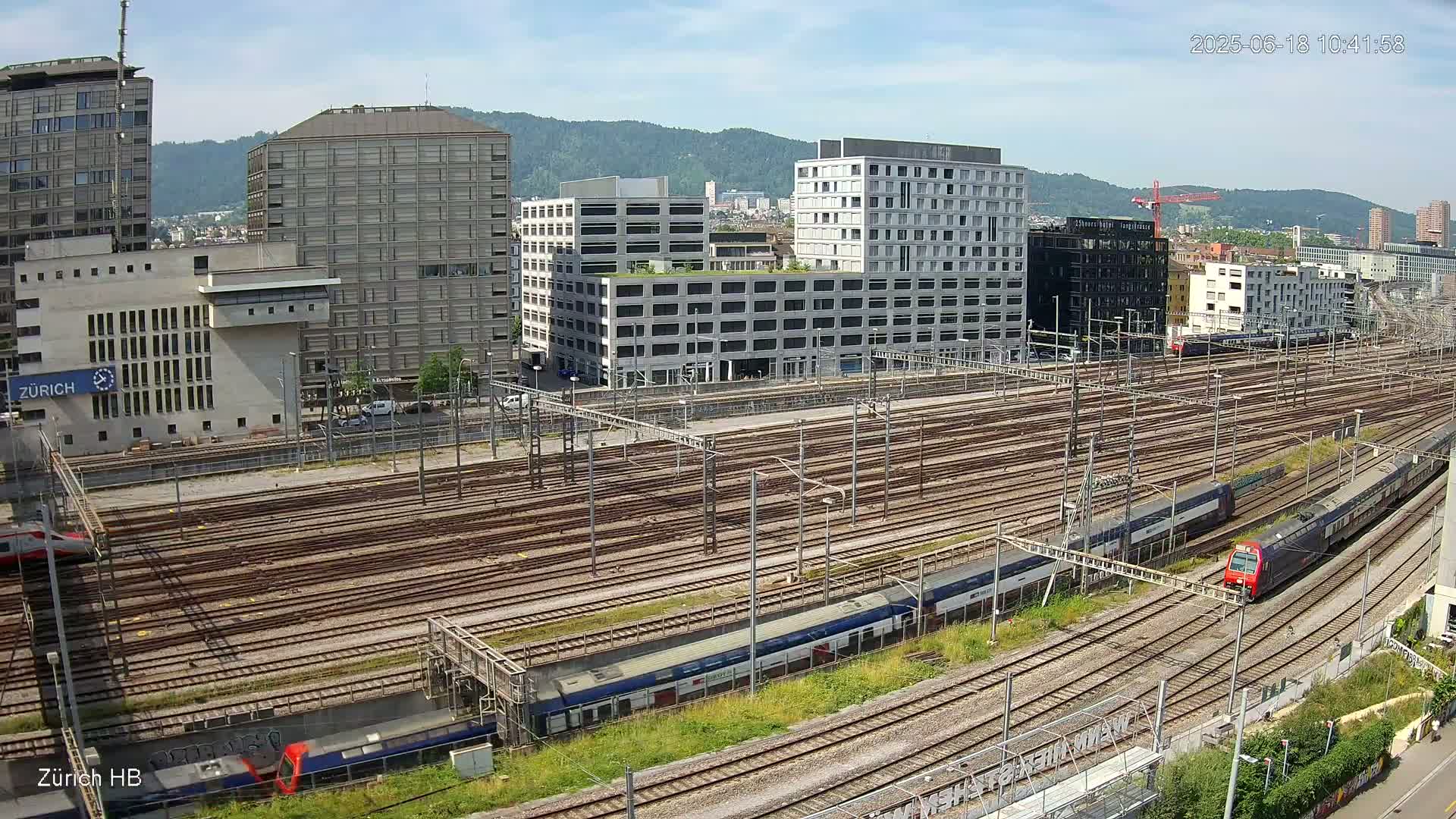 A sunny day view of a large train yard surrounded by modern buildings and mountains in the background.