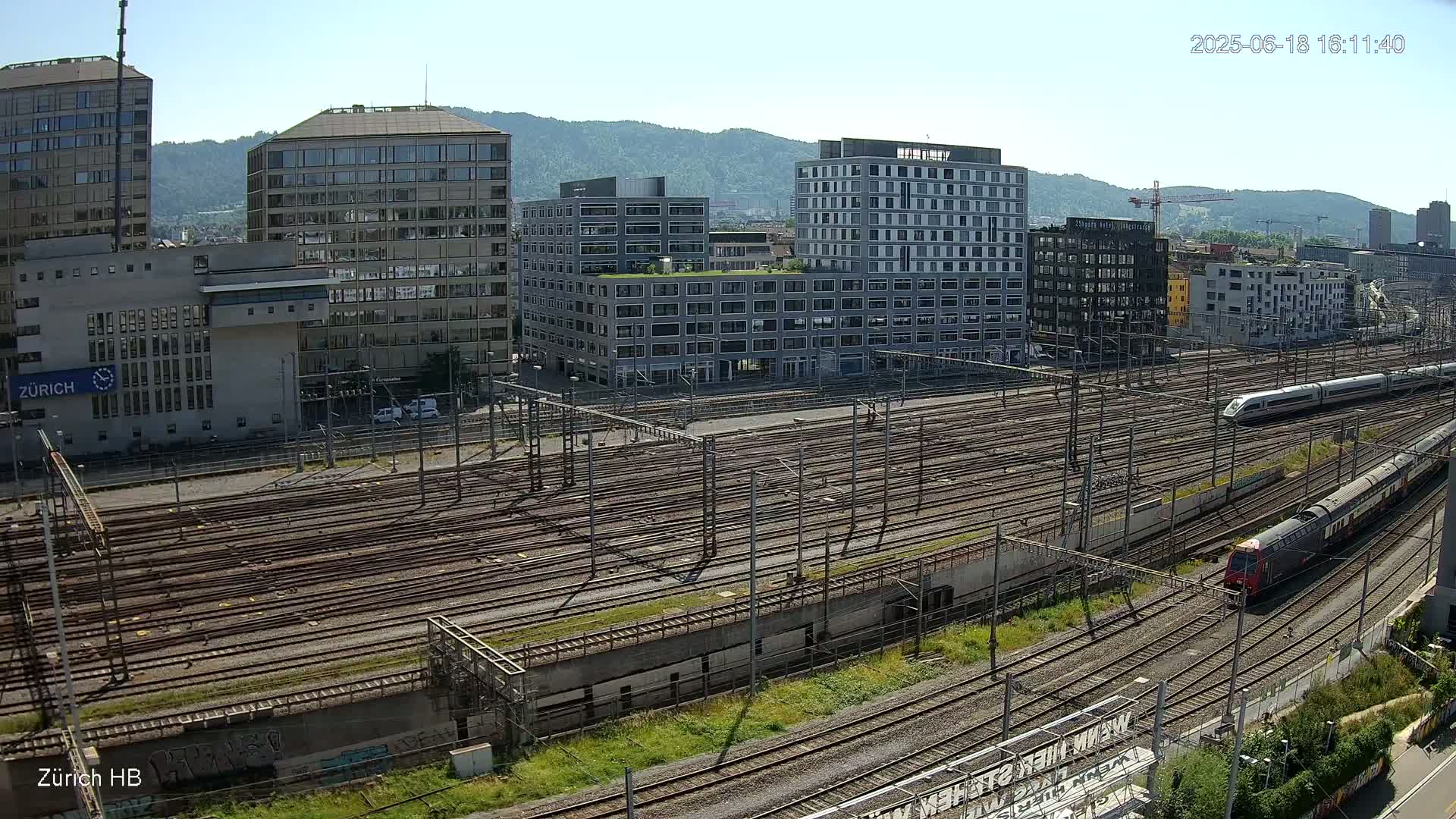 A high-angle, sunny view of a large train yard with multiple tracks, surrounded by modern buildings and mountains in the background.