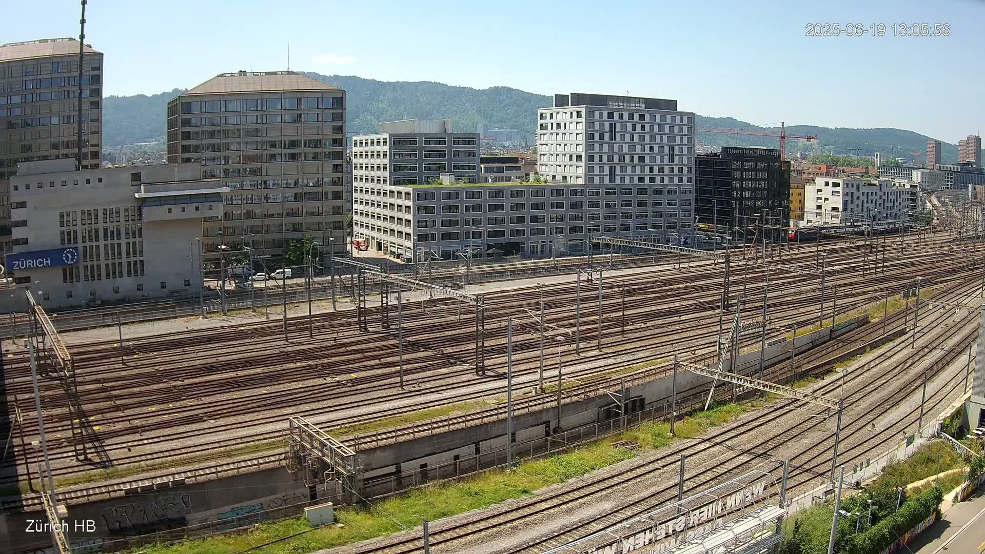 A sprawling network of train tracks stretches out beneath a sunny sky, surrounded by modern buildings and hills in the distance.