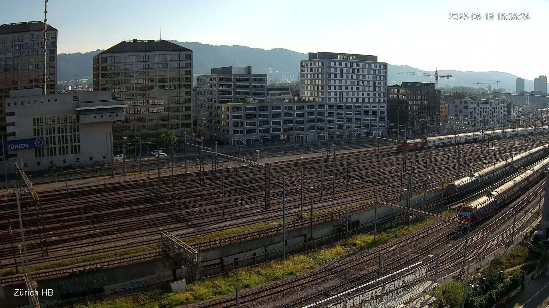 A high-angle, long shot of a train station on a sunny day, showing numerous train tracks with several trains present, alongside several modern buildings and hills in the background.
