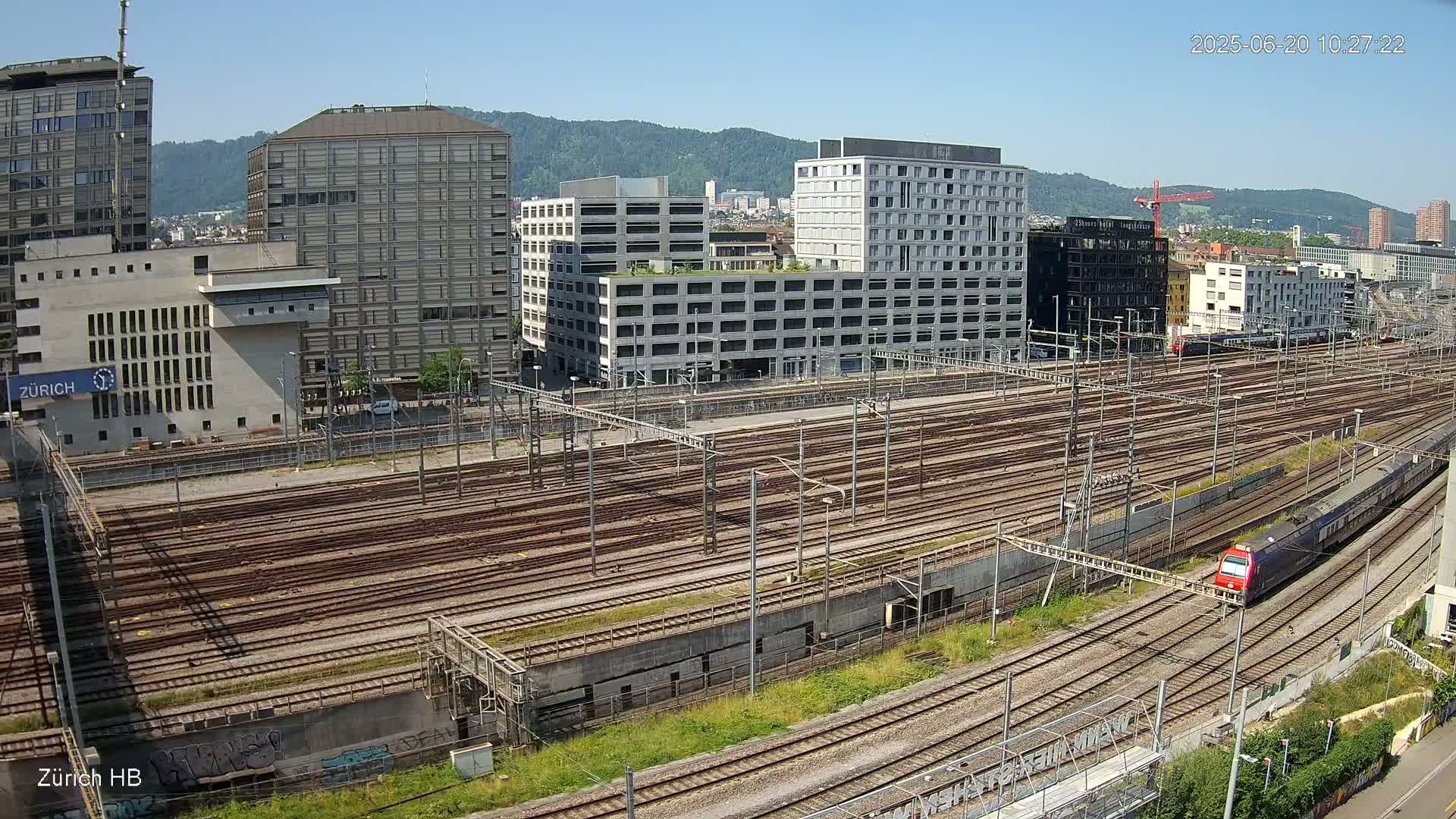A high-angle, long shot on a sunny day shows a sprawling train yard filled with numerous parallel train tracks, surrounded by modern buildings and a distant mountain range.