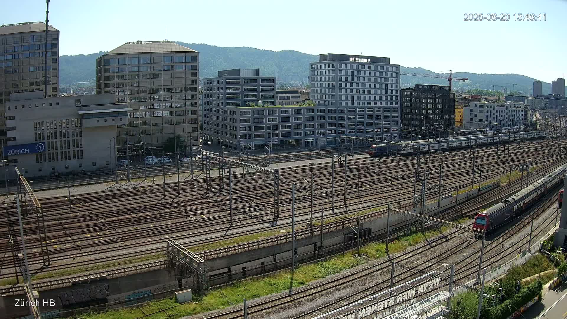 A sunny day reveals a sprawling train yard with numerous tracks, surrounded by modern and older buildings, including high-rises and low-rise structures, with trains visible on the tracks.