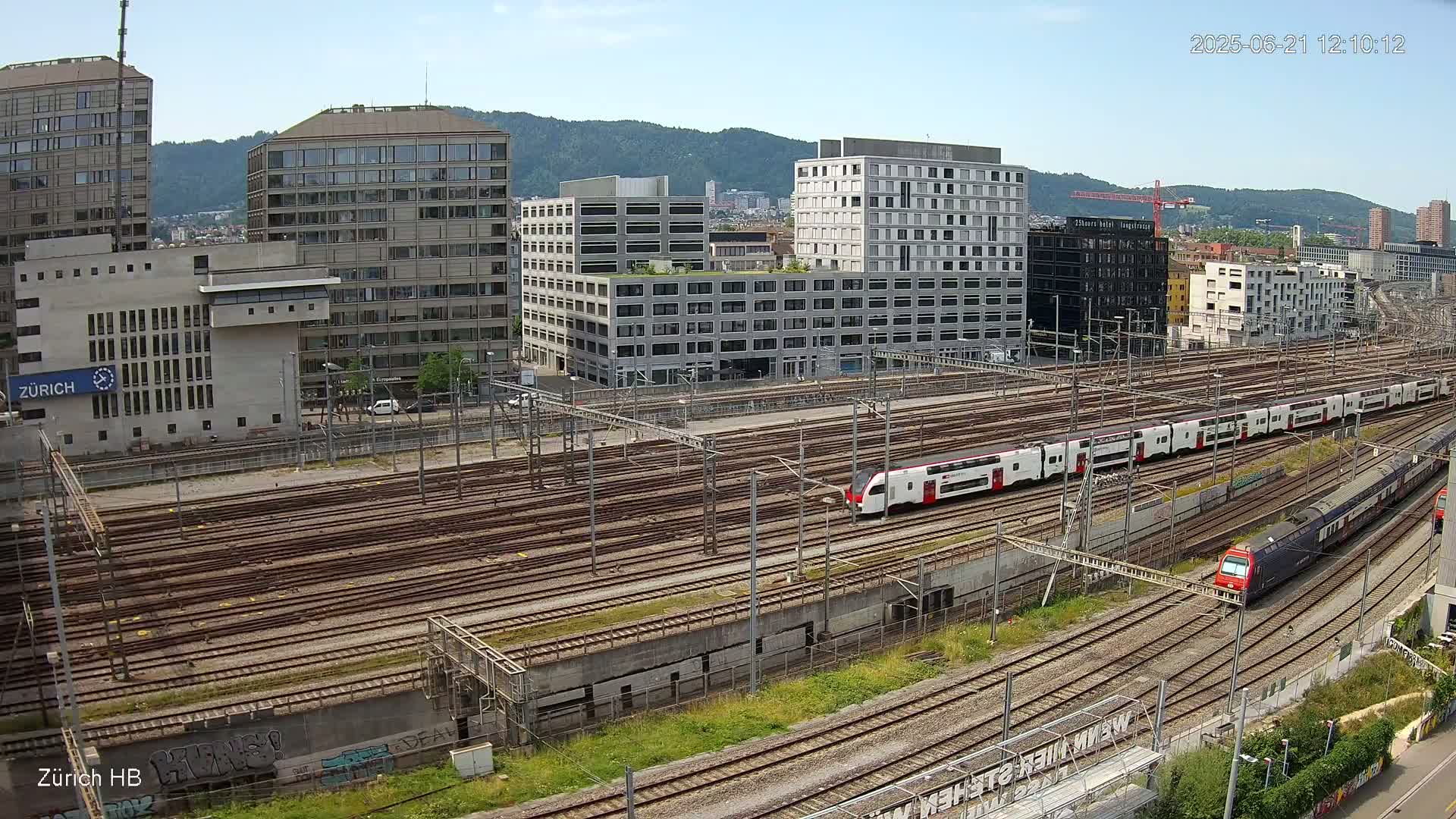 A high-angle view of a train station on a sunny day with several trains on numerous tracks, surrounded by modern buildings and a mountain in the background.