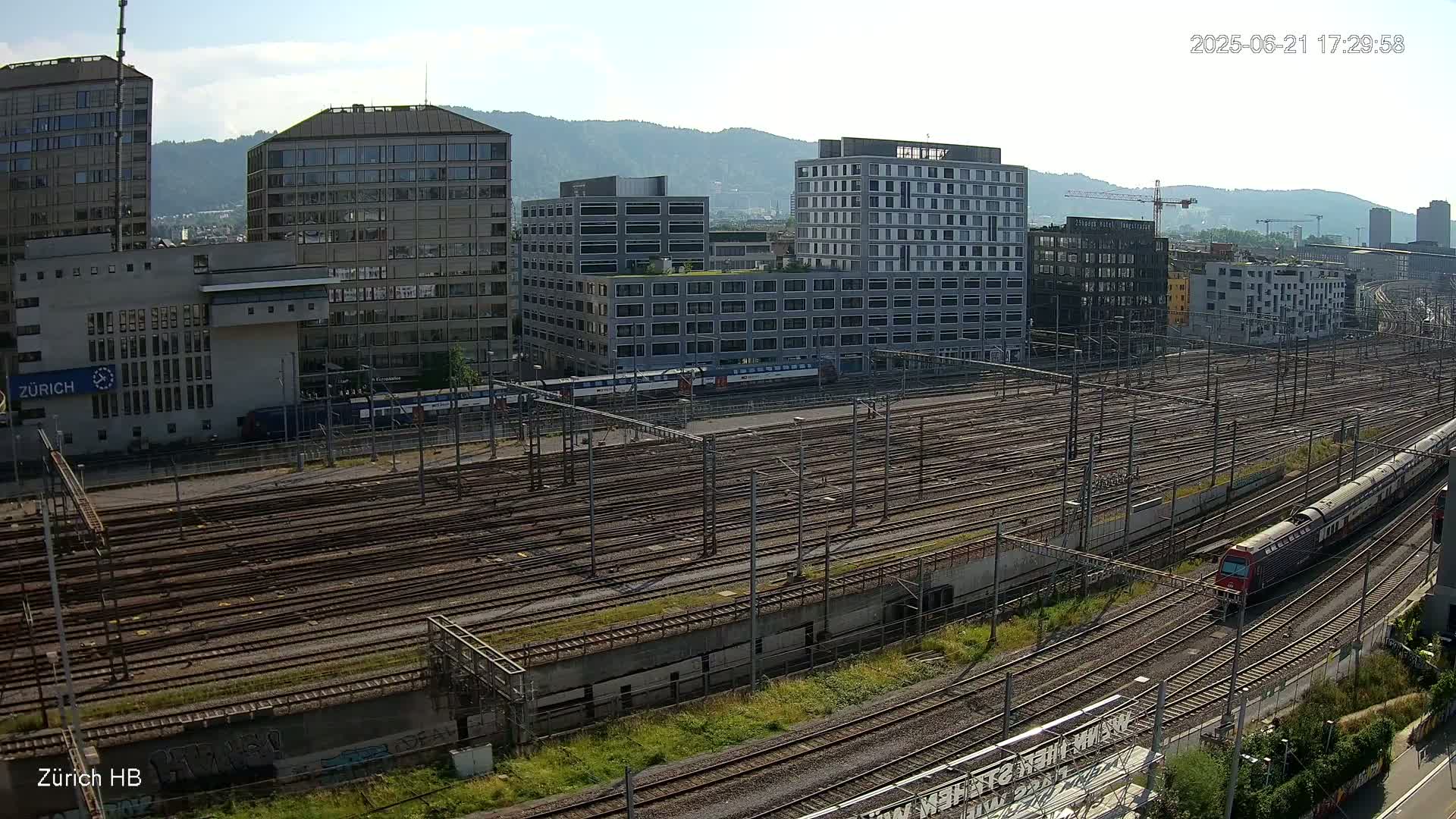 A sunny day reveals a sprawling railway yard surrounded by modern buildings and mountains in the distance, with several trains visible on the tracks.