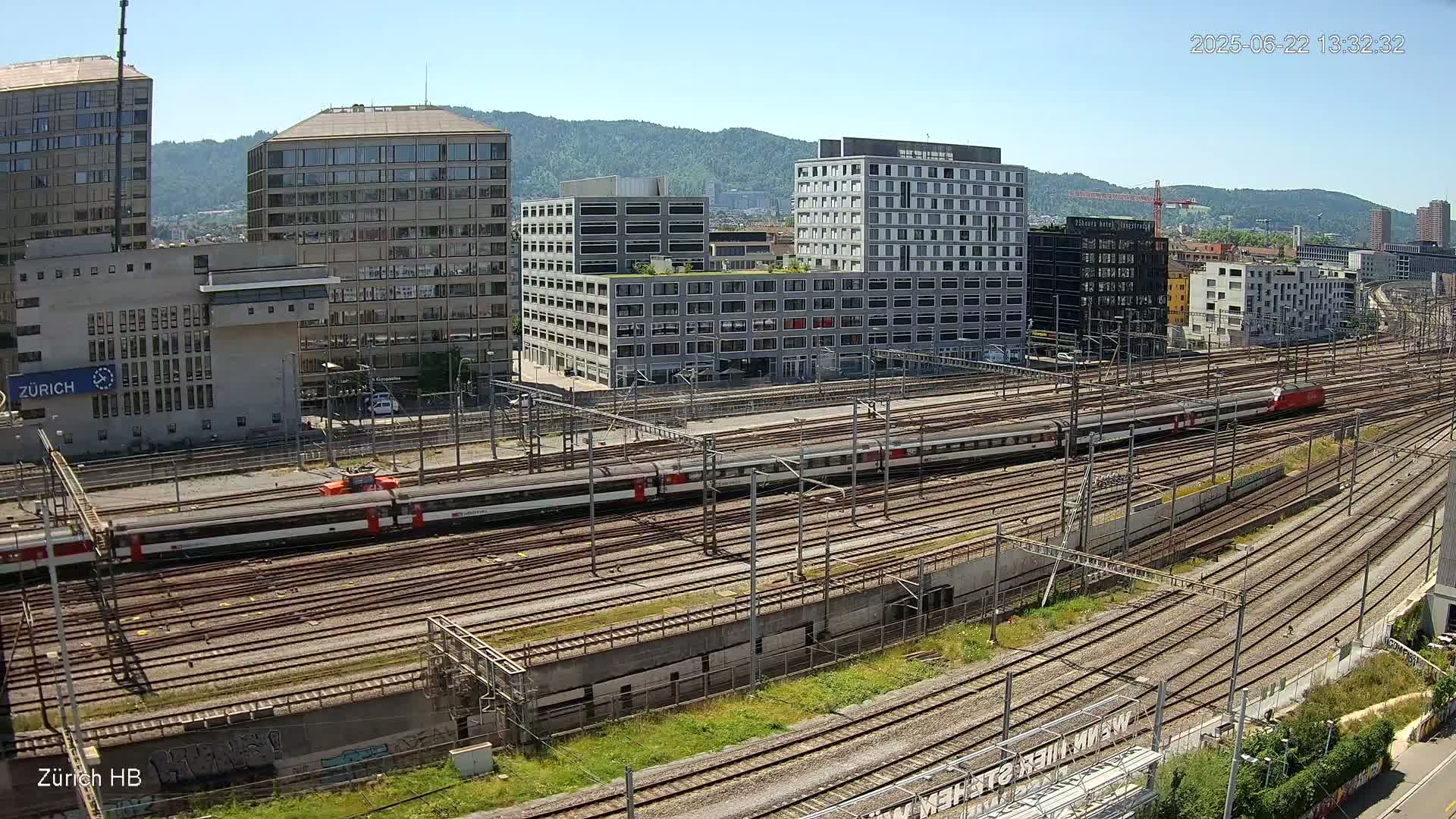 A sunny day shows a sprawling railway yard with multiple train tracks and several modern buildings in the background.