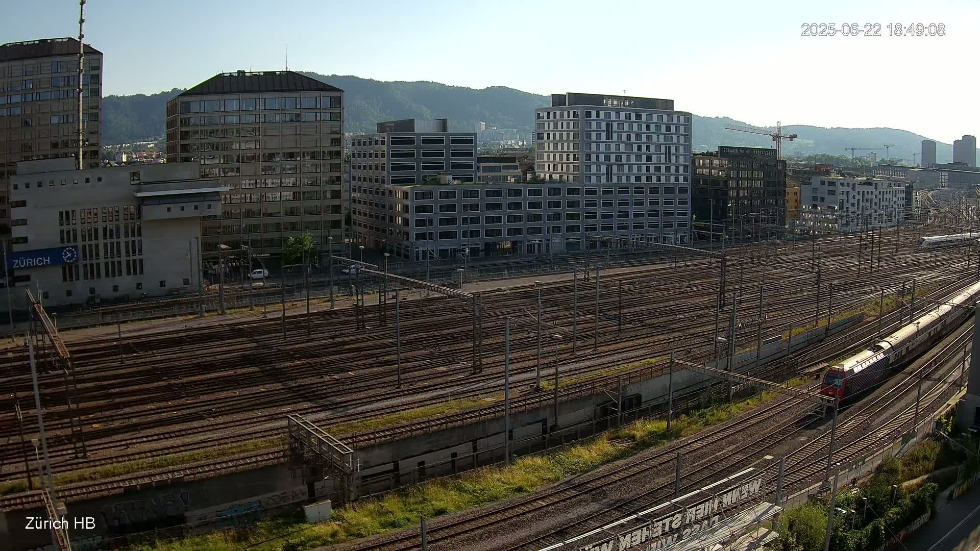 A high-angle, long shot shows a train traveling on a vast network of train tracks next to several modern buildings on a sunny day with mountains in the background.