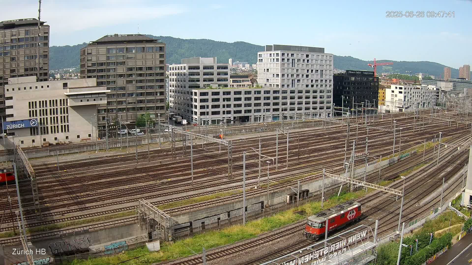 A red locomotive travels across a large network of train tracks in front of several modern buildings on a partly sunny day.