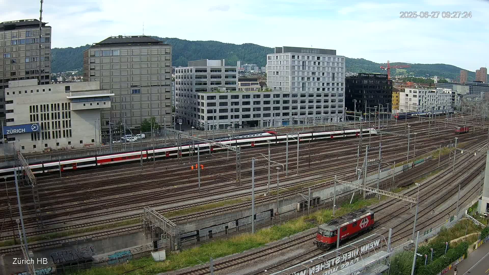 A long passenger train sits on a network of train tracks surrounded by modern buildings under a partly cloudy sky.