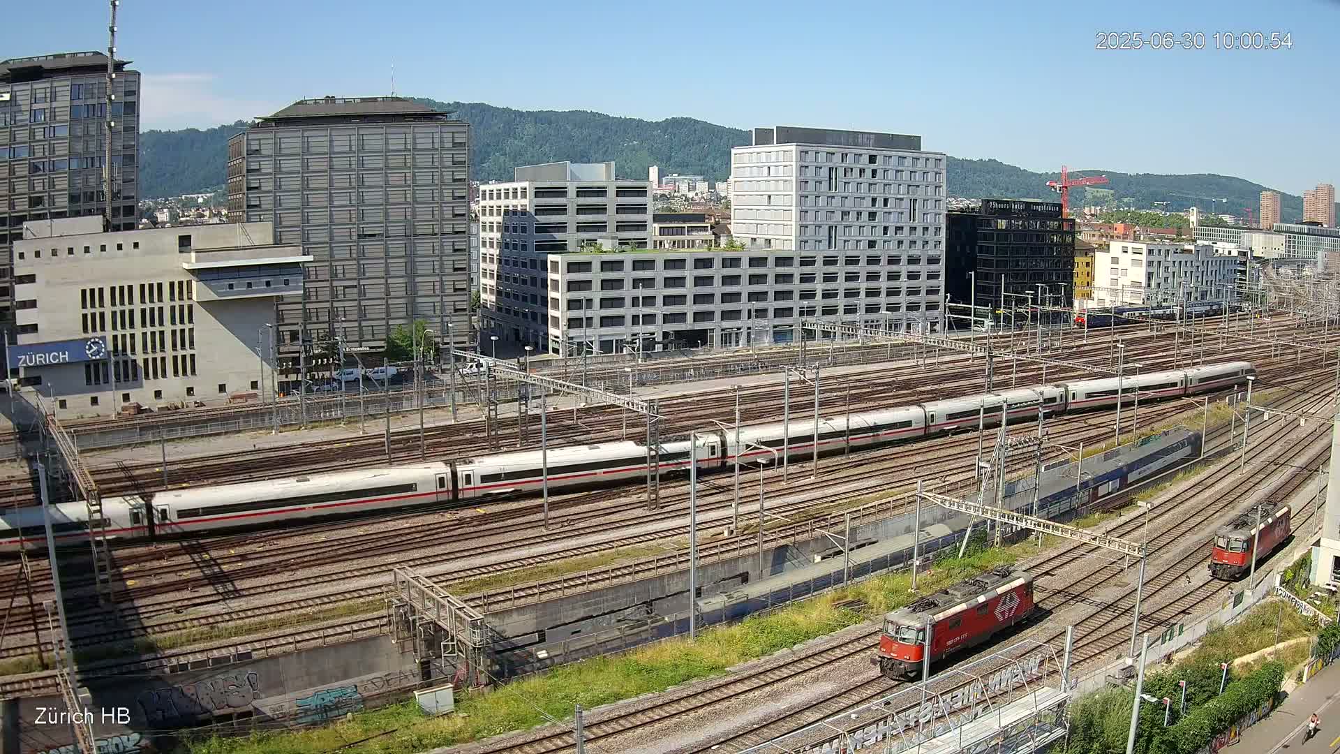 A high-angle, long shot on a sunny day shows a large railway yard with multiple trains, including a long high-speed train, and several buildings in the background.