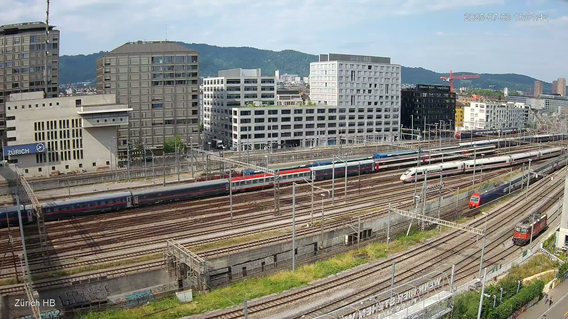 A high-angle view of a large railway yard with multiple trains on sunny day, surrounded by modern buildings and hills in the background.