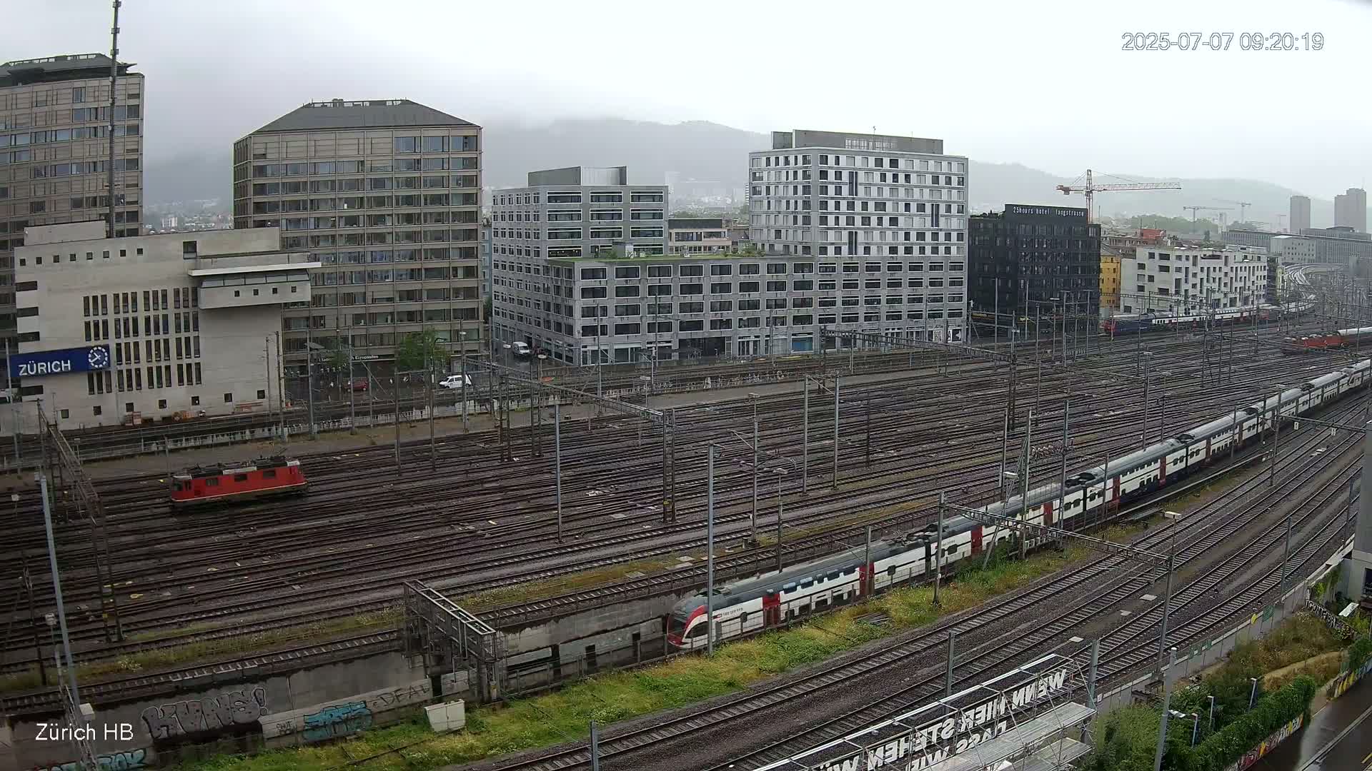 A long passenger train and a smaller red locomotive are visible on a sprawling network of train tracks next to several multi-story buildings in rainy weather.