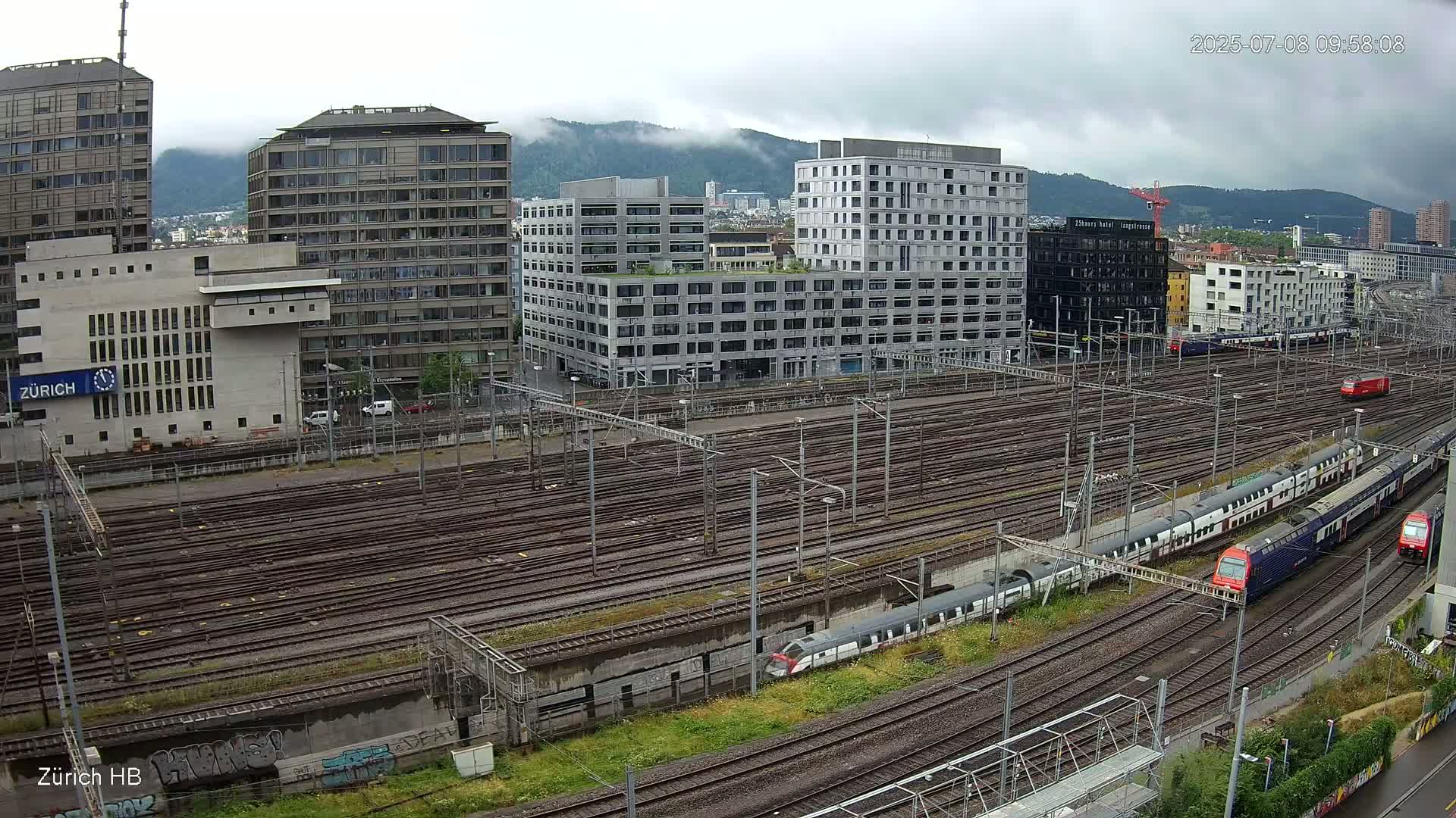A sprawling railway yard with numerous tracks, surrounded by modern buildings under an overcast sky.