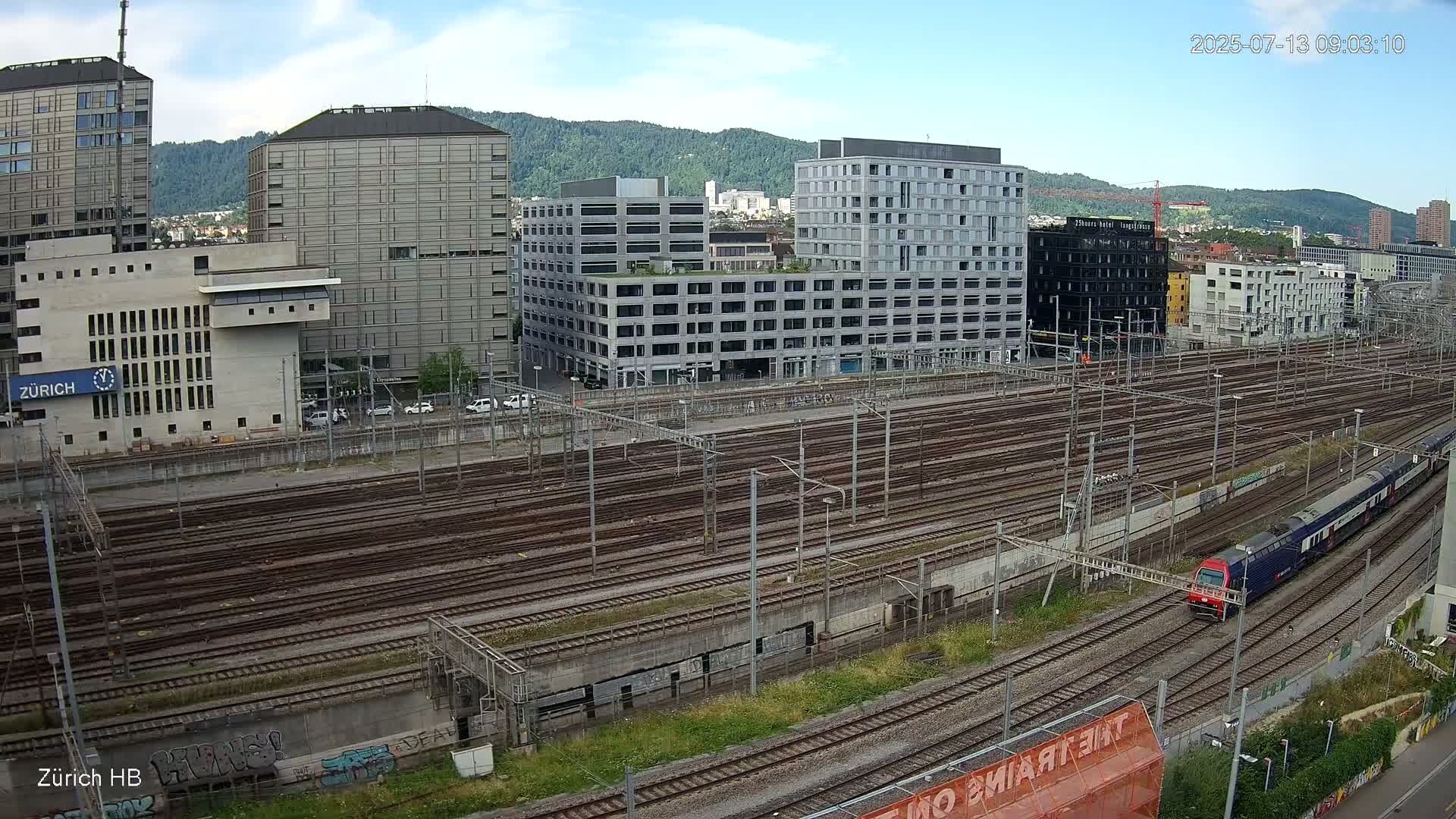 A red and white passenger train travels along numerous railway tracks next to several multi-story buildings under a partly sunny sky.