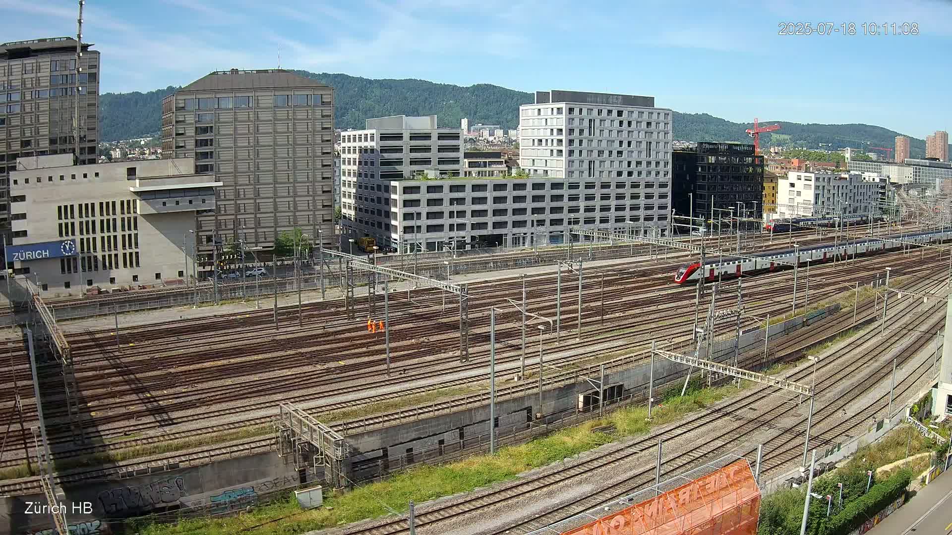 A sunny day overlooking a sprawling railway yard with numerous train tracks, surrounded by modern buildings and hills in the background, with a train visible on one of the tracks.