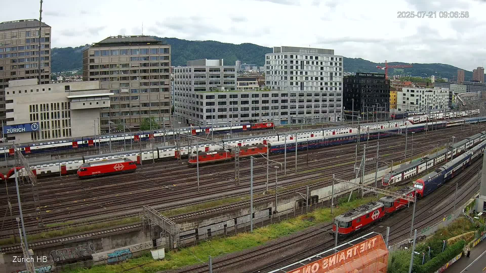 A high-angle view shows numerous trains on a railway network next to several multi-story buildings under an overcast sky.