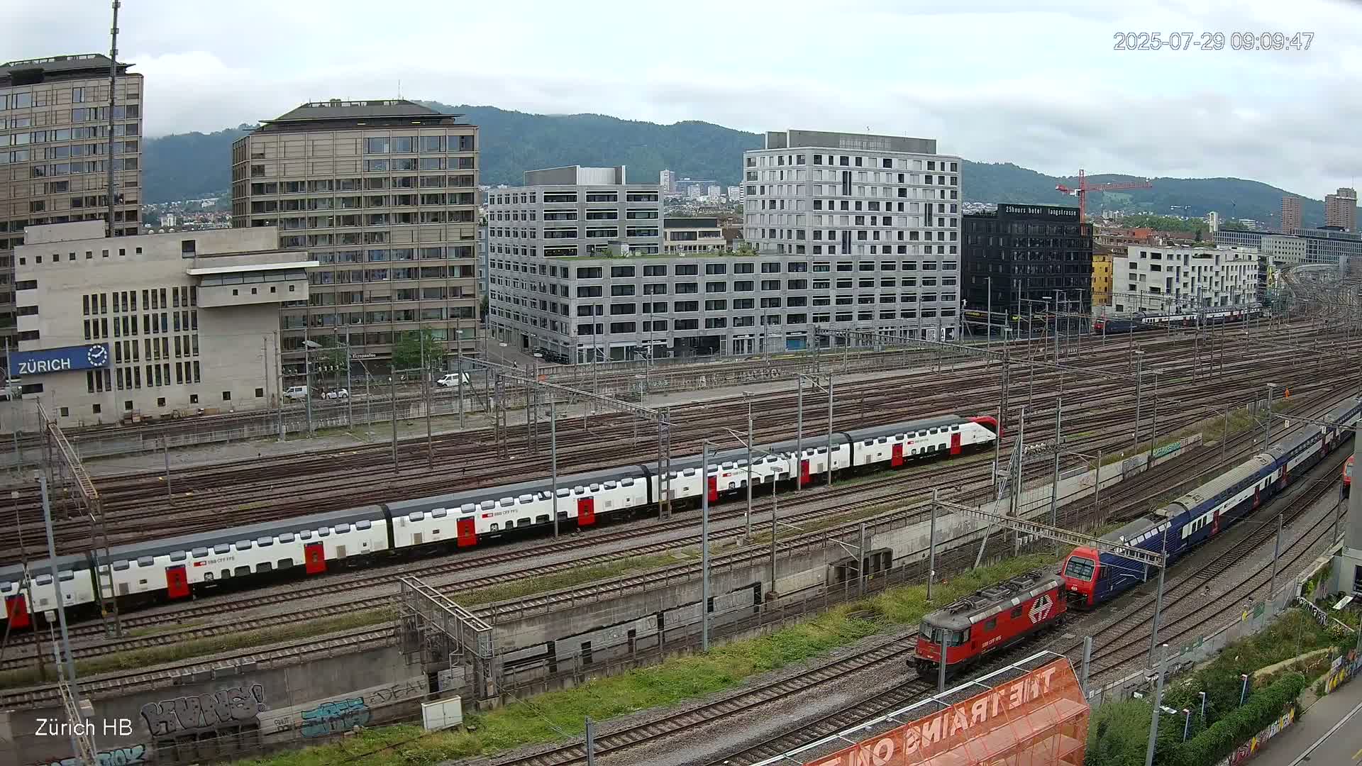 Two passenger trains and a locomotive are visible on a sprawling railway network next to several modern buildings under an overcast sky.