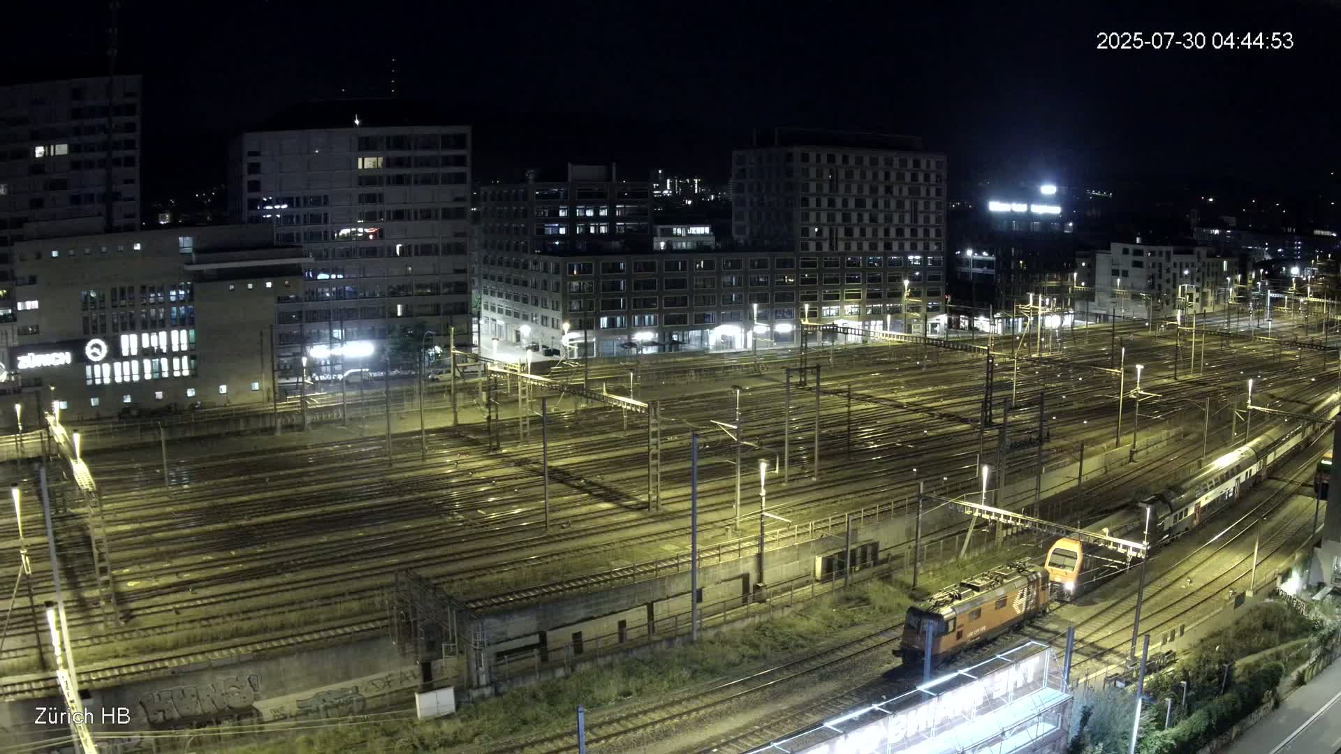 Two passenger trains and a locomotive are visible on a sprawling railway network next to several modern buildings under an overcast sky.