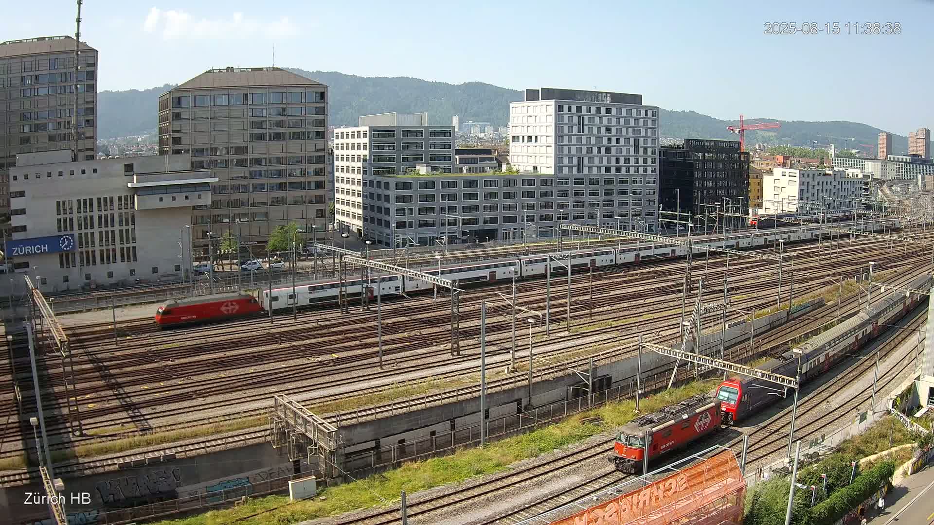 Two passenger trains and a locomotive are visible on a sprawling railway network next to several modern buildings under an overcast sky.