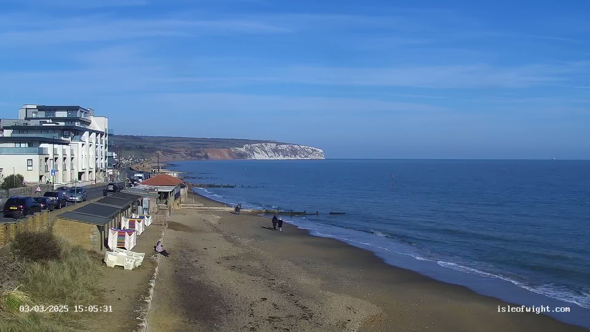 A sandy beach with a few people walking, beach huts, and a building line the shore, overlooking a calm ocean and distant white cliffs under a clear blue sky.