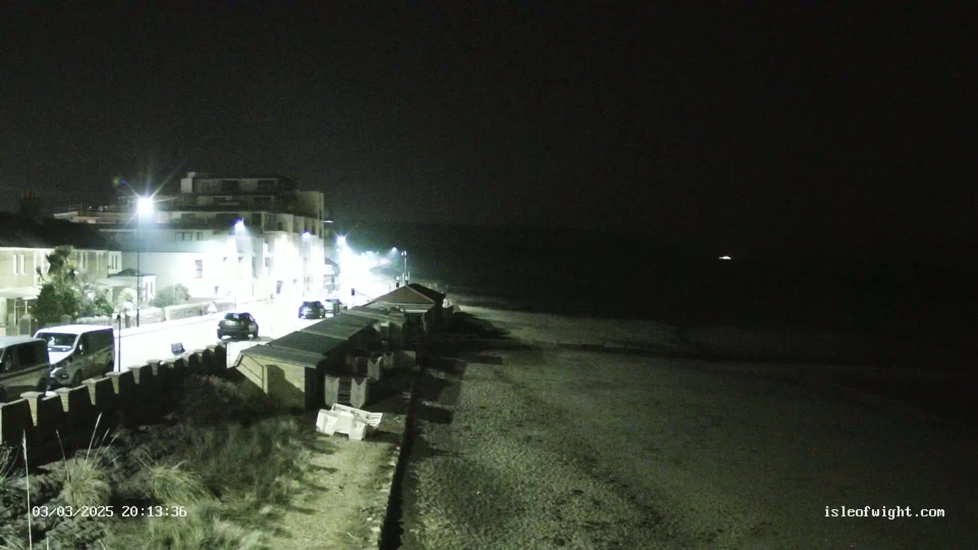 A nighttime view of a seaside town with a street of buildings, parked cars, beach huts, and a dark, pebbled beach extending to a dark ocean under a clear sky.