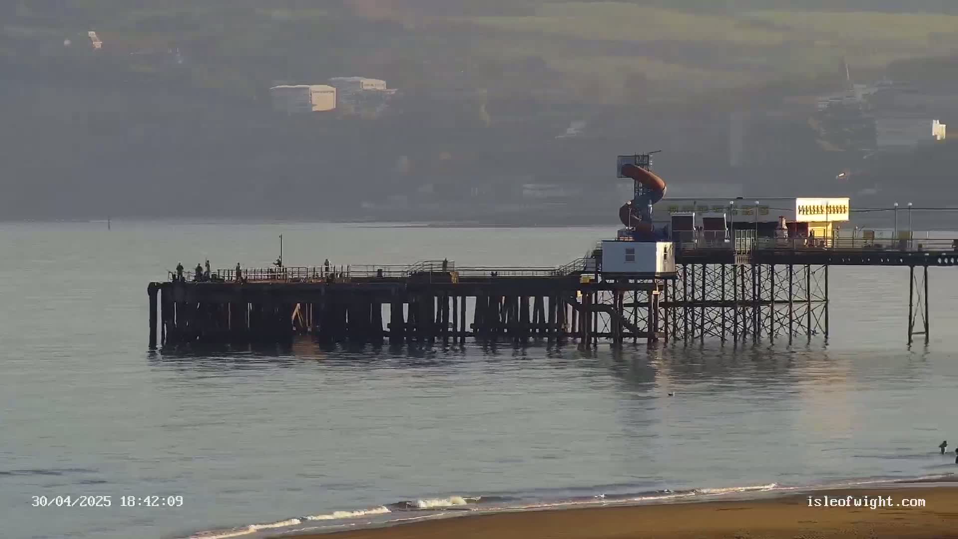 A long pier extends into a calm, gray sea under a hazy sky, with a few people visible on the pier and a sandy beach in the foreground.
