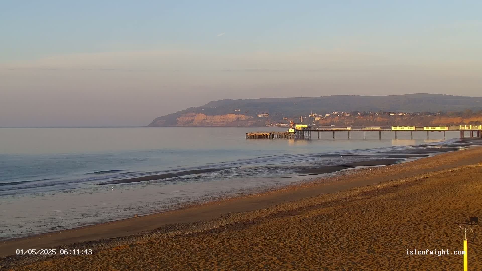 A long sandy beach stretches toward a pier extending into a calm sea under a pale sky, with cliffs visible in the distance.
