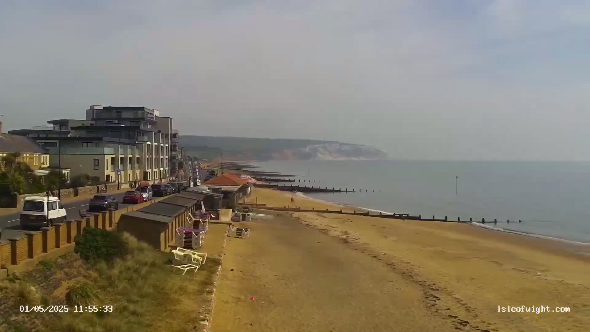 A hazy day reveals a sandy beach, a row of beach huts, buildings along a road, and distant cliffs overlooking a calm sea.