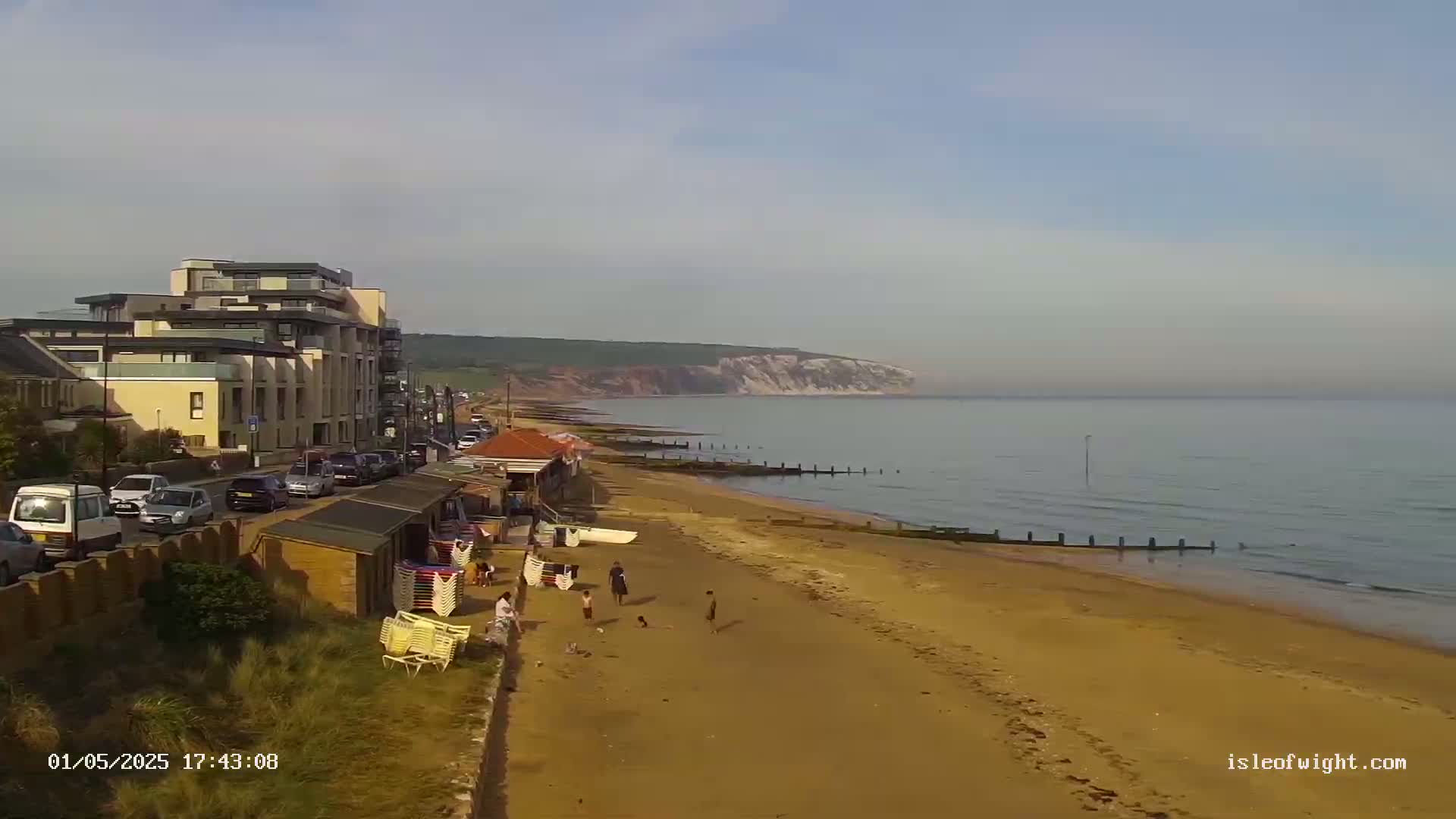 A sandy beach with a few people and beach chairs stretches towards a calm ocean and white cliffs under a clear sky.