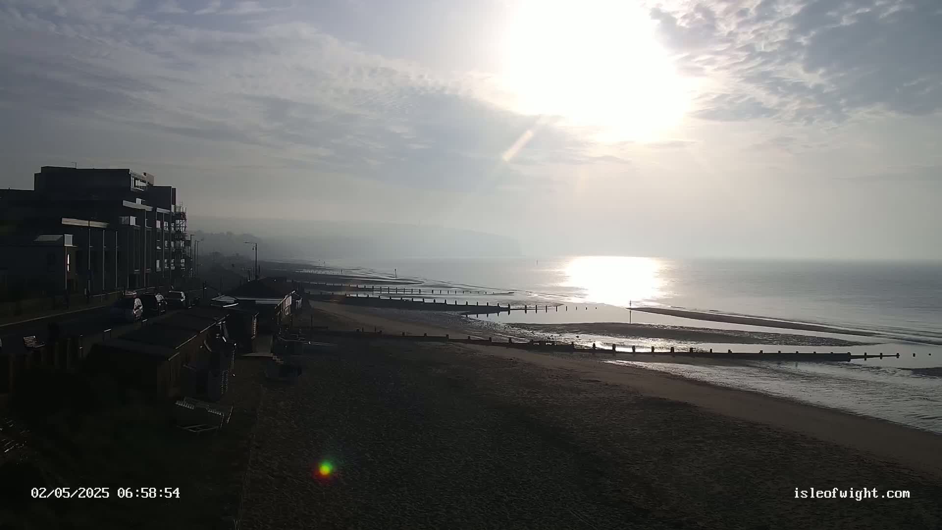 A hazy sunrise over a calm sea illuminates a beach with wooden barriers, viewed from a seaside town with buildings and parked cars.