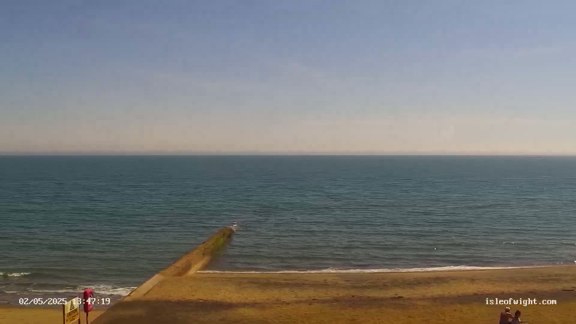 A calm, hazy sea meets a sandy beach, where two people are visible in the lower-right corner.