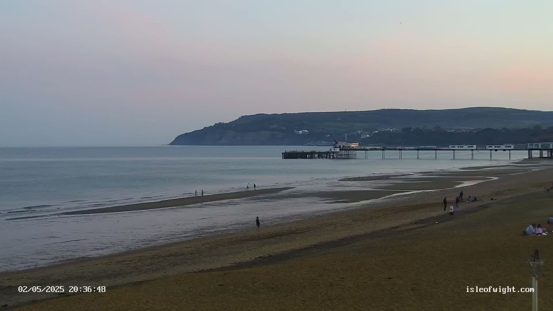 A long pier extends into a calm sea at dusk, with a sandy beach and a few people visible, beneath a hill in the background.