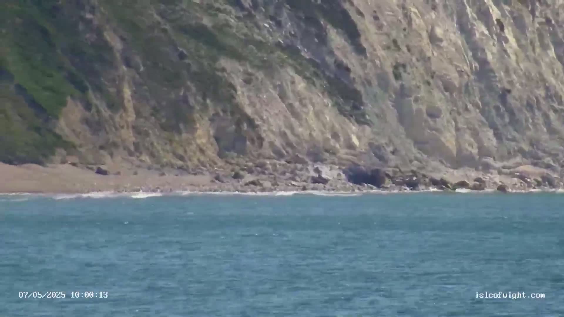A rocky cliff overlooking a calm, blue ocean with a small, sandy beach at its base.