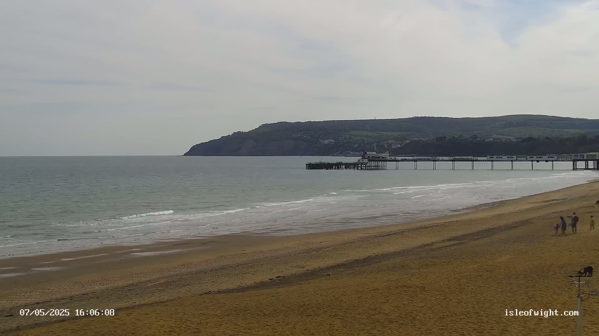 A sandy beach stretches toward a calm sea with small waves, a long pier extending into the water, and a hill in the background under a mostly cloudy sky.