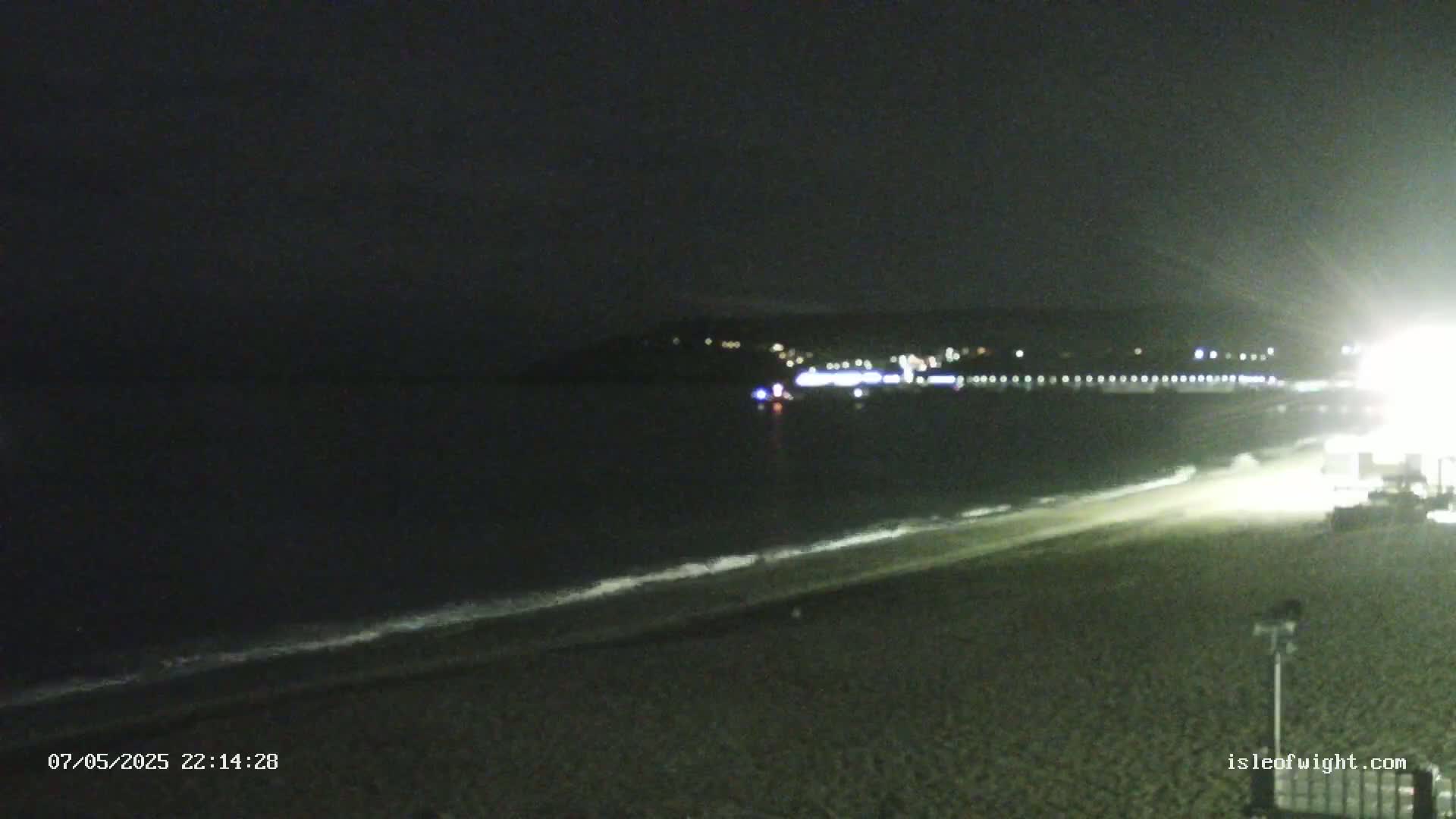 A dark, nighttime beach scene shows a calm ocean with a distant, faintly lit shoreline.