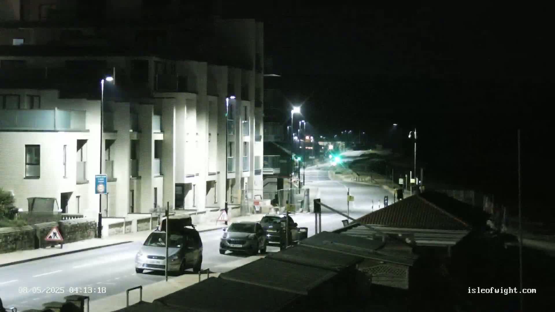 At night, a mostly empty road illuminated by streetlights passes in front of a modern apartment building and leads toward a dimly lit beach.