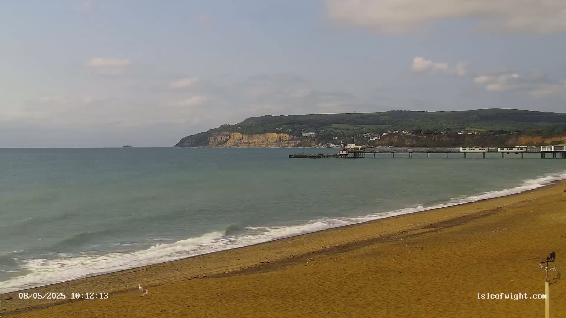A sandy beach with gentle waves laps the shore, a pier extends into a calm sea toward a distant cliff, under a partly cloudy sky.
