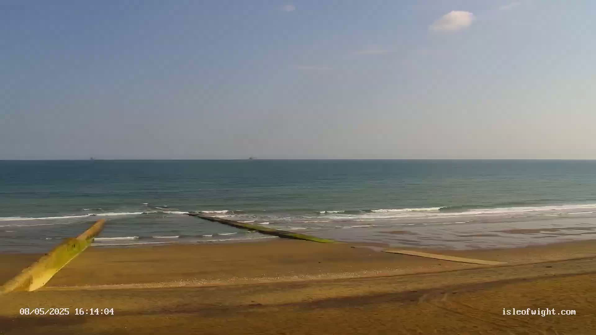 A sandy beach with calm ocean waves and two long, low concrete structures extending from the shore into the water under a mostly clear sky.