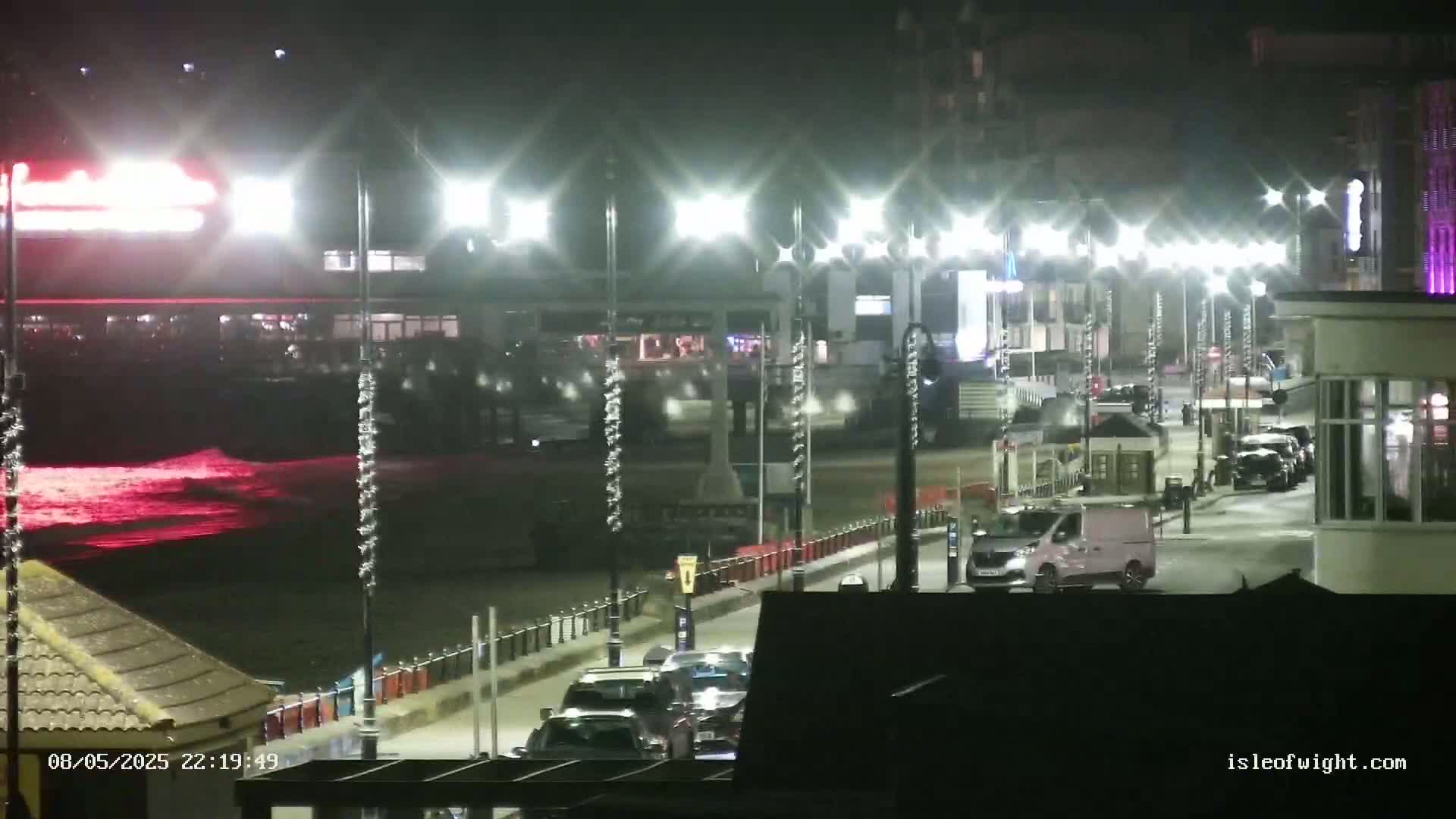 A nighttime scene of a seaside promenade with brightly lit buildings, several cars, and a van, overlooking a dark ocean with red-tinged waves.
