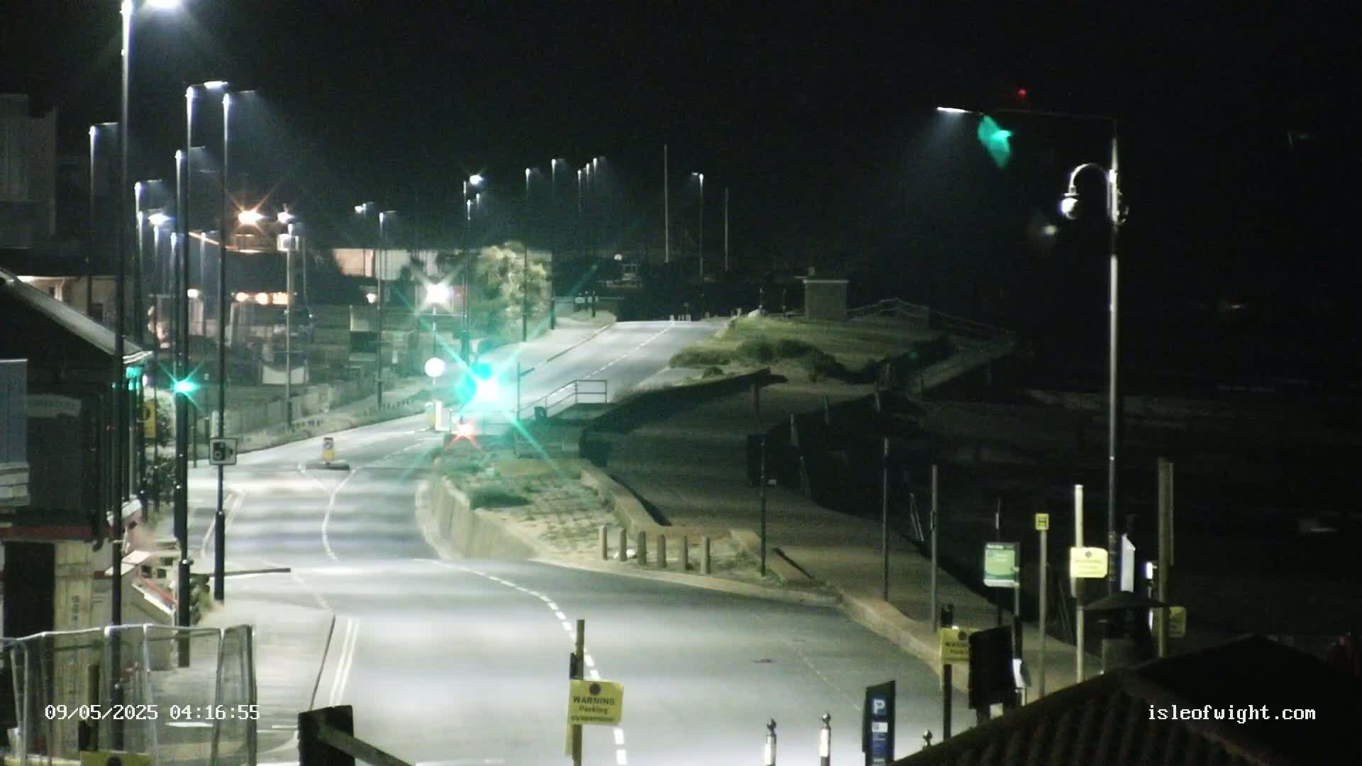 A nighttime, brightly lit roadway curves gently downward toward a shoreline, with several streetlights illuminating the scene.