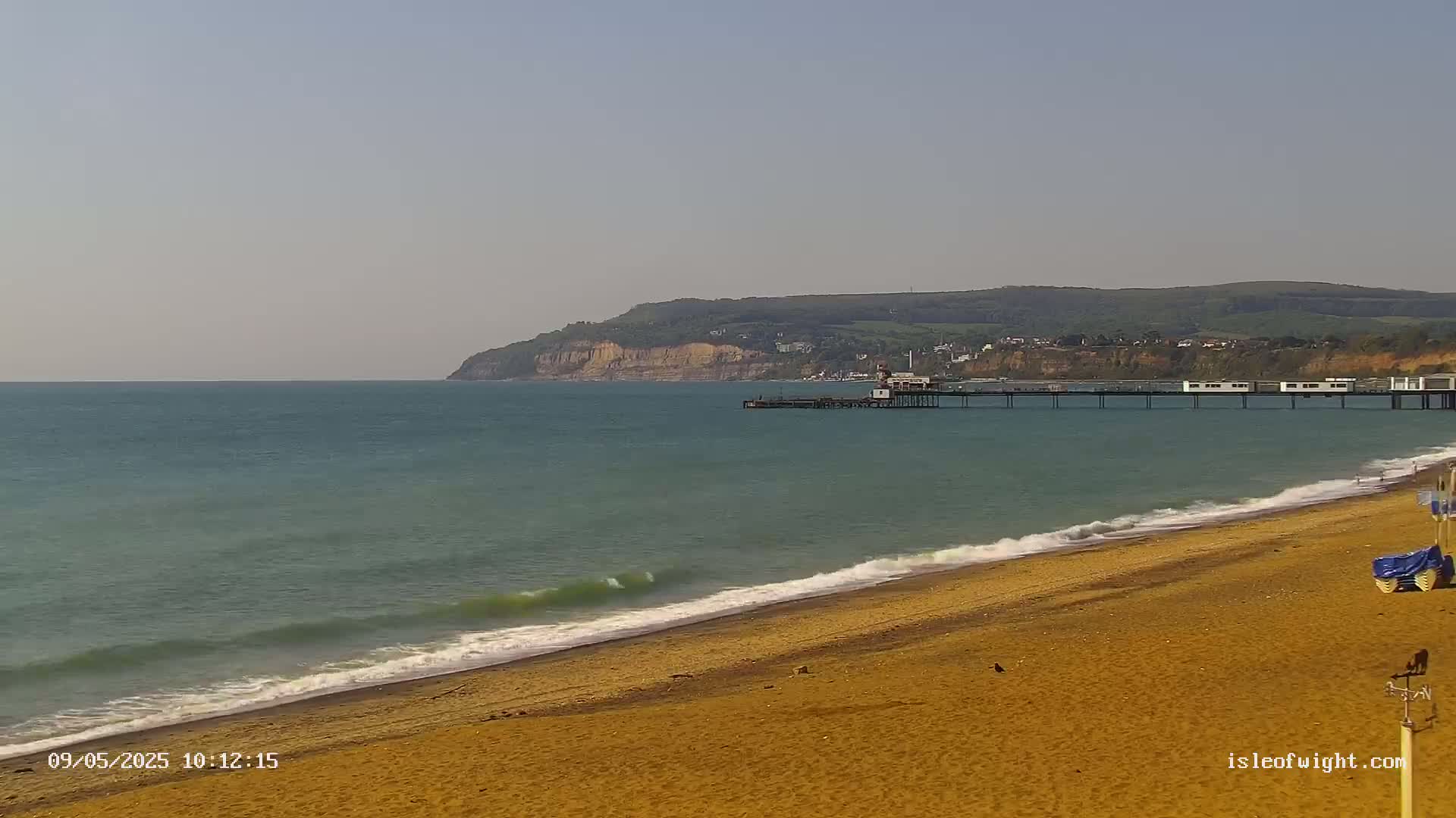 A sandy beach with gentle waves laps the shore, a pier extends into a calm sea towards a distant cliff face under a clear sky.
