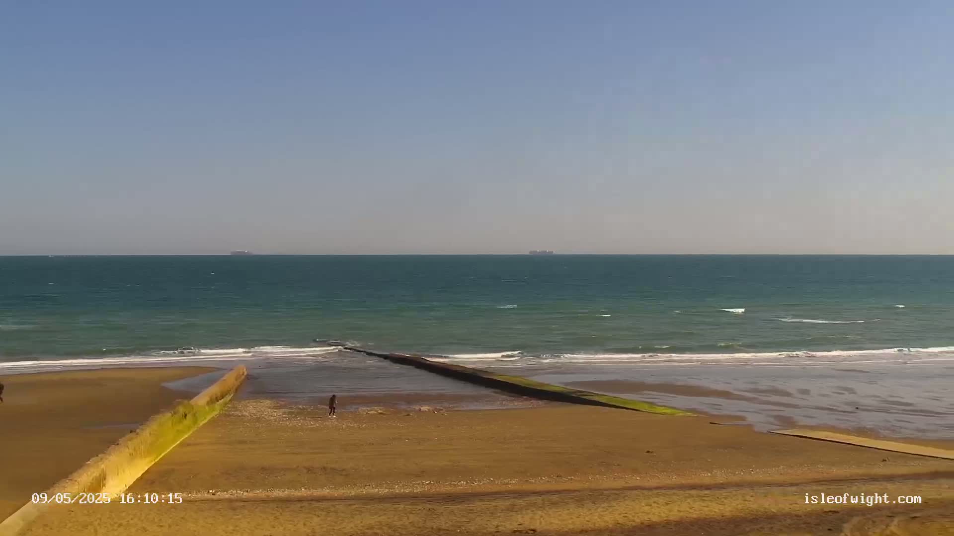 A sandy beach slopes down to a calm, teal ocean under a clear blue sky, with a long, thin structure extending from the beach into the water and a few people visible in the distance.