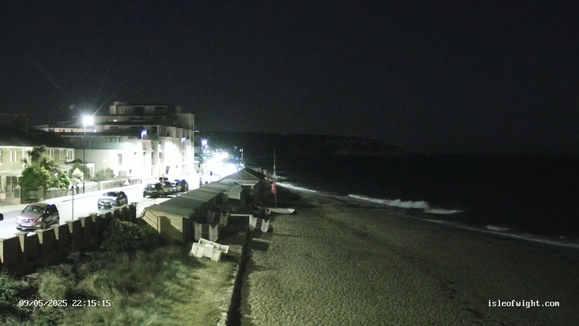A nighttime view shows a beach with gentle waves, a row of beach huts, a roadway with parked cars, and a building behind it, all under a dark sky.