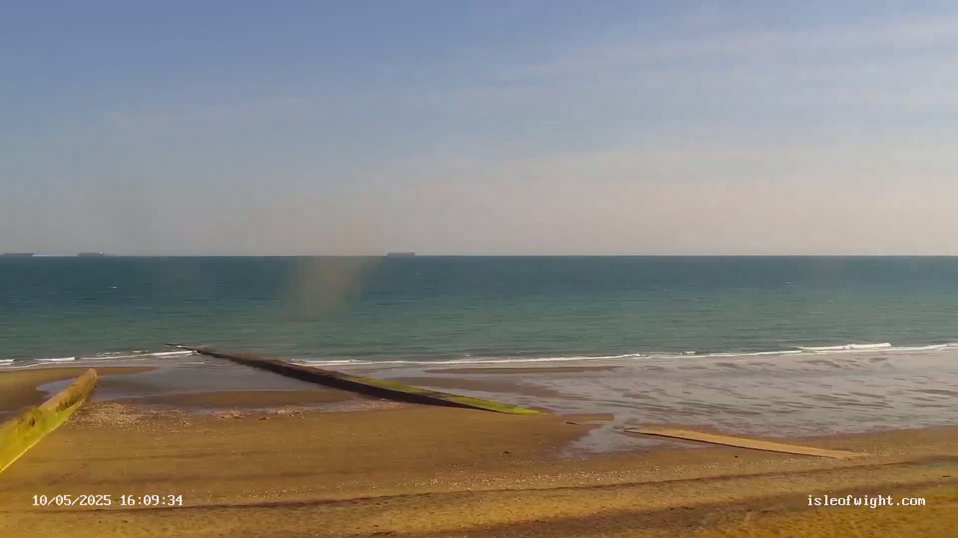A sandy beach slopes down to a calm, teal ocean under a clear, bright sky, with a man-made structure leading from the land into the water.