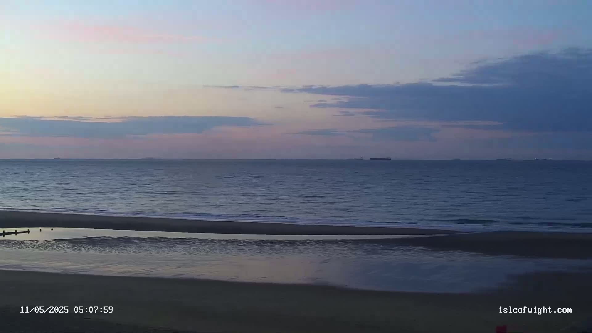 A calm ocean meets a sandy beach under a soft, pastel sunrise sky with a few clouds.