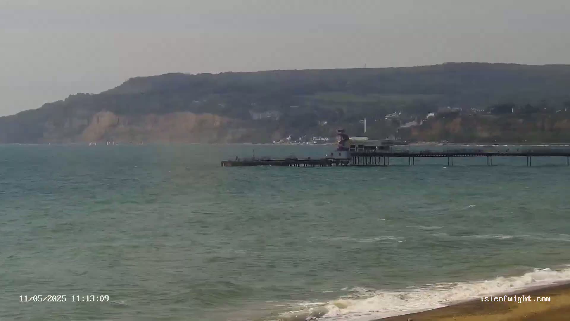 A long pier extends into a choppy sea, with a hilly coastline and a few windsurfers visible in the distance under an overcast sky.