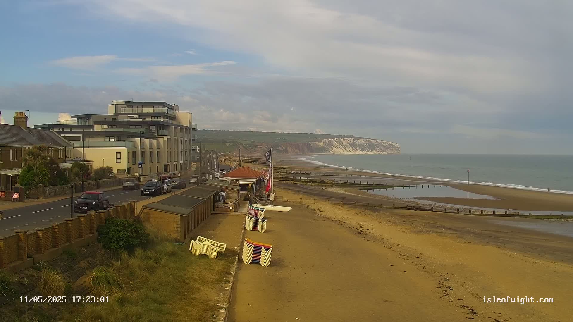 A sandy beach with stacked beach chairs, buildings, and cars, stretches towards a calm ocean and white cliffs under a mostly cloudy sky.